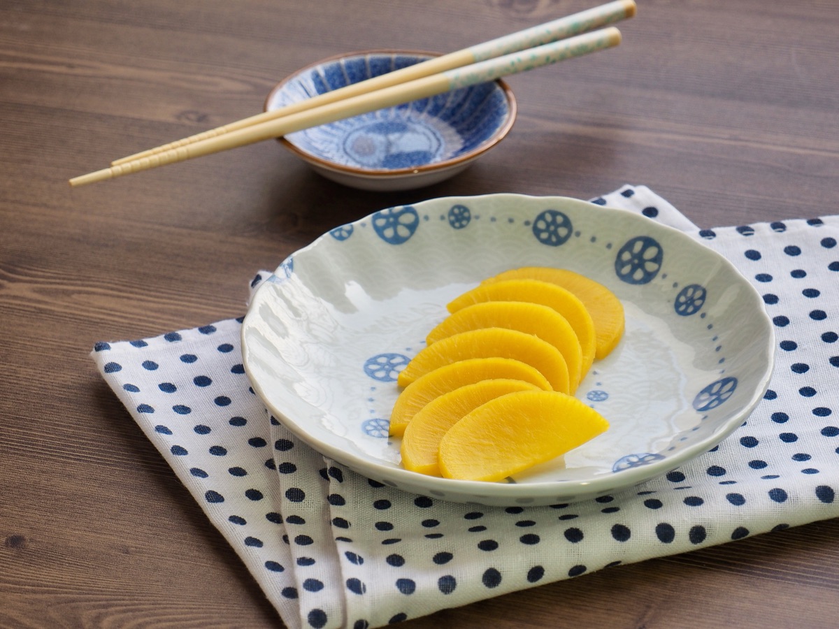 Rondelles de radis jaune coréen (danmuji) disposées sur une assiette en céramique bleue, avec des baguettes et un petit bol de sauce sur une table en bois.