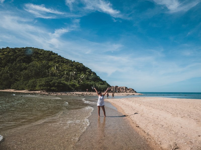 Maud levant les bras sur une plage en Thaïlande, entre mer turquoise et colline verdoyante